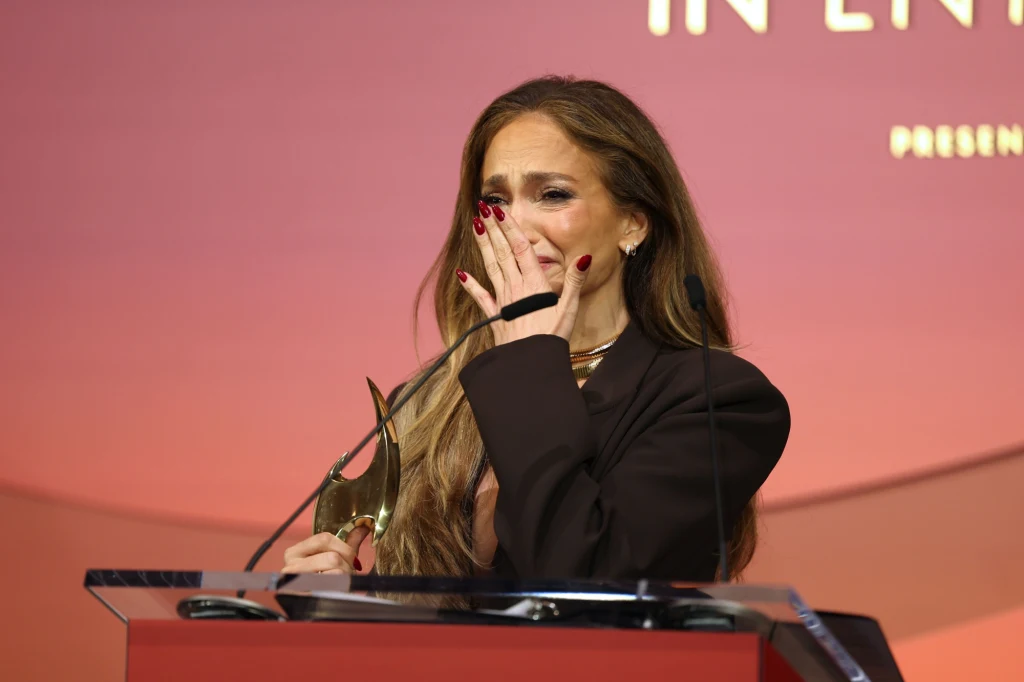 Kobieta stojąca przy mównicy z mikrofonem, emocjonalnie zasłaniająca usta dłonią, w drugiej ręce trzyma statuetkę. Tło w ciepłych odcieniach różu z rozmytymi napisami.