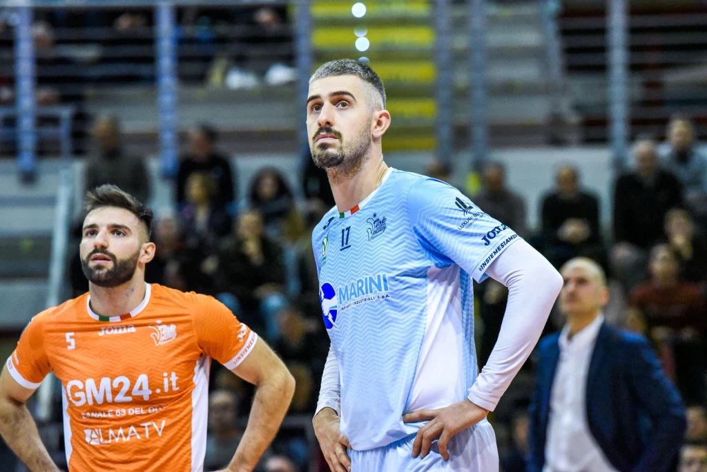 Michele Baranowicz Dwóch siatkarzy stojących na boisku podczas meczu, jeden w niebieskim stroju, drugi w pomarańczowym, w tle widownia na trybunach.