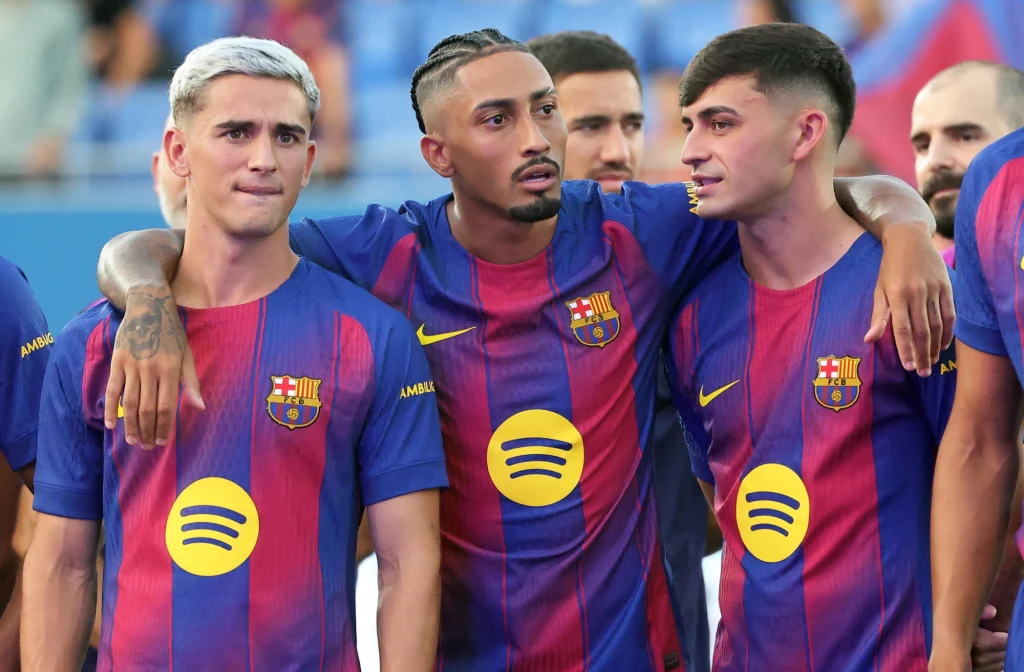 Trzech młodych piłkarzy w koszulkach FC Barcelony, stojących obok siebie na tle trybun stadionowych. Dwóch z nich obejmuje się nawzajem, wszyscy są skupieni i patrzą w różne strony.