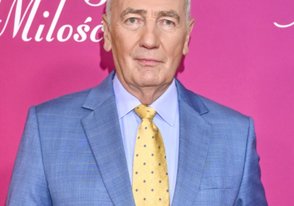 Starszy mężczyzna w niebieskiej marynarce i żółtym krawacie na tle różowej ściany z białym napisem.