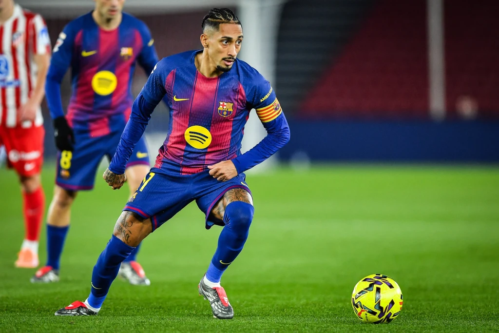 Piłkarz w stroju FC Barcelony z opaską kapitańską prowadzi piłkę podczas meczu na zielonej murawie, w tle widoczni inni zawodnicy i stadionowe trybuny.