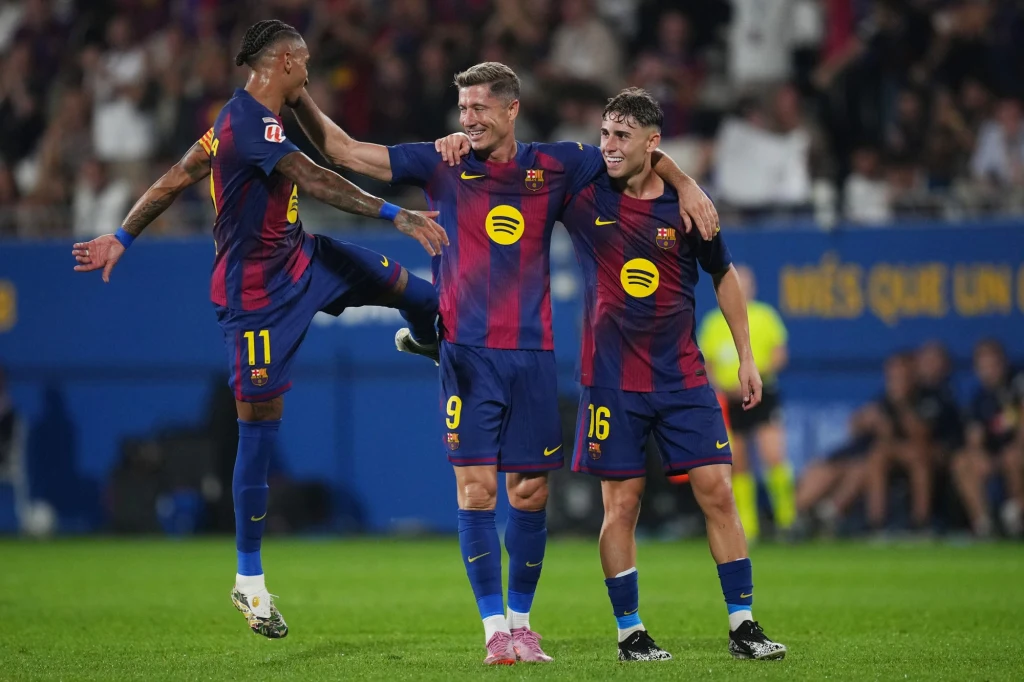 Trzech piłkarzy w granatowo-bordowych strojach FC Barcelony świętuje razem na boisku, jeden zawodnik podskakuje z uniesioną nogą, a pozostali uśmiechają się i obejmują. Tło rozmyte, widownia i reflektory.