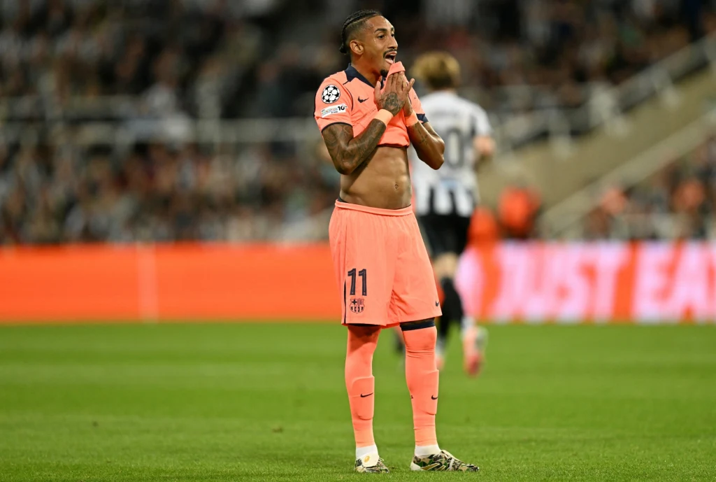 Piłkarz ubrany w różowy strój sportowy z numerem 11 stoi na murawie stadionu, podciąga koszulkę do góry pokazując fragment brzucha, w tle rozmazane postaci innych zawodników oraz trybuny z kibicami.