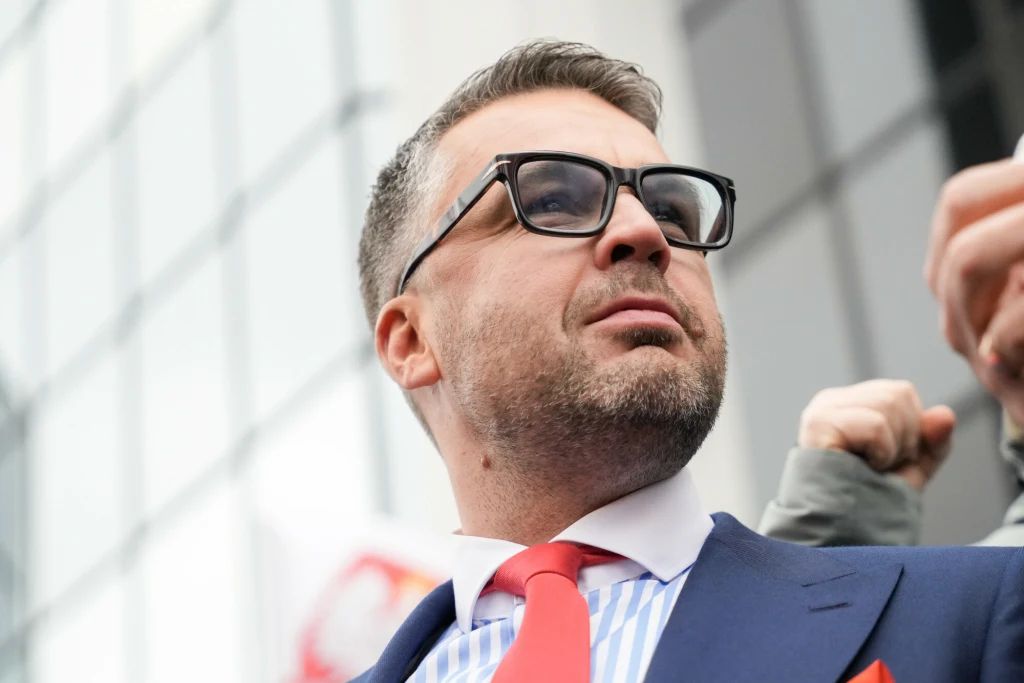 Mężczyzna w garniturze i czerwonym krawacie z brodą i okularami przeciwsłonecznymi stoi na tle nowoczesnego budynku ze szkła, prezentując postawę pewności siebie.