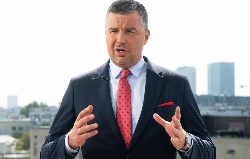 Mężczyzna w garniturze z czerwonym krawatem stoi na tle panoramy miasta z widocznymi wieżowcami, gestykulując podczas wypowiedzi.