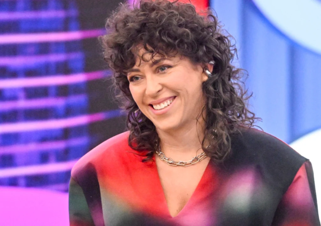 Uśmiechnięta kobieta z kręconymi, ciemnymi włosami w kolorowej marynarce, nosząca srebrny łańcuszek. W tle widoczne rozmyte elementy scenografii telewizyjnej.