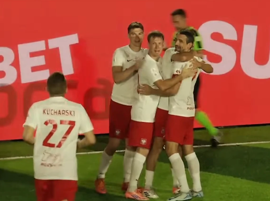 Grupa piłkarzy w białych koszulkach i czerwonych spodenkach świętuje zdobycie gola, przytulając się na tle dużego czerwonego ekranu z białym napisem, jeden z zawodników z numerem 27 odwrócony plecami.