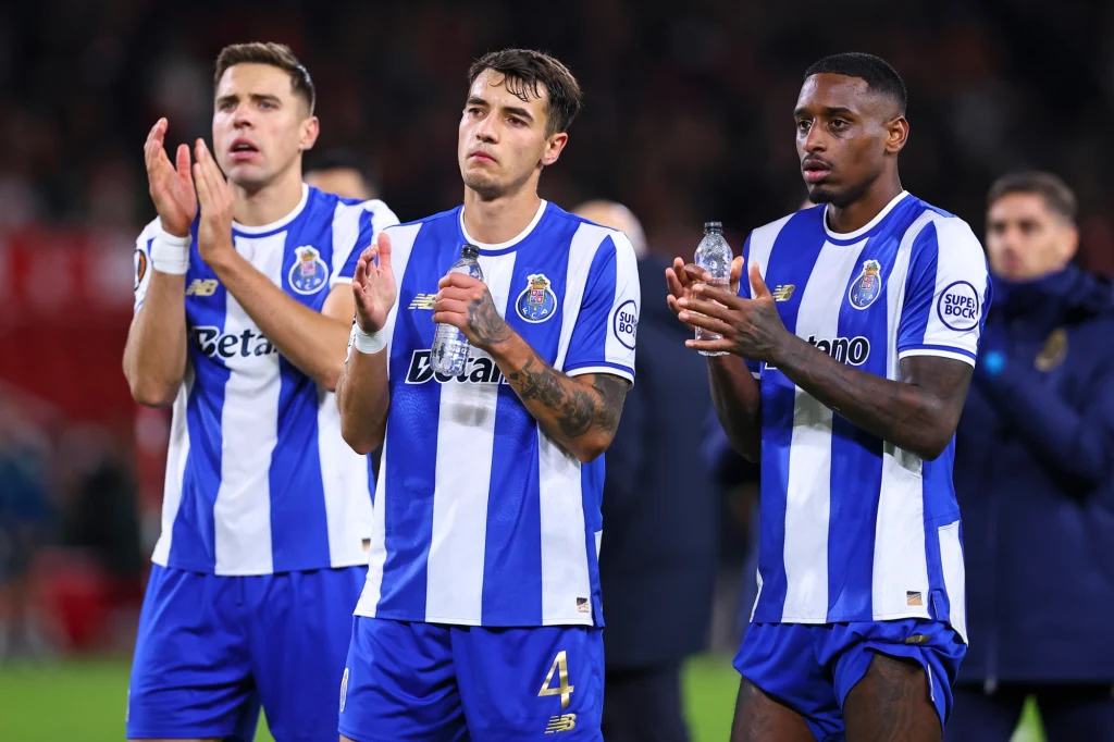 Trzech piłkarzy drużyny FC Porto w biało-niebieskich strojach stoi na boisku klaskając, trzymają w rękach plastikowe butelki z wodą, tło rozmyte z innymi osobami.
