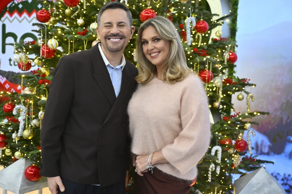 Uśmiechnięty mężczyzna i kobieta pozują razem przed dużą, udekorowaną choinką z kolorowymi bombkami, światełkami oraz wstążkami, tworząc świąteczną i radosną atmosferę.