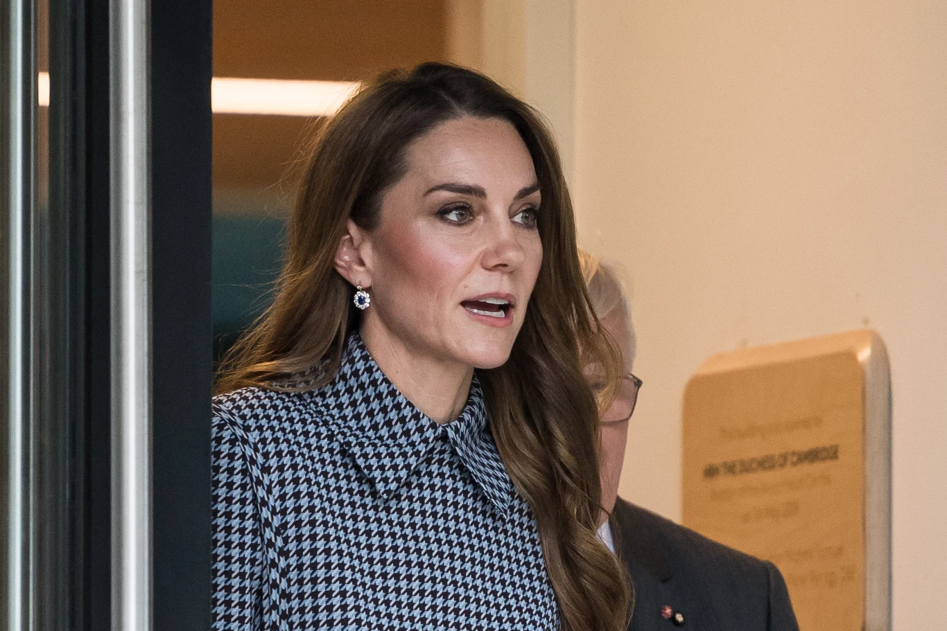 Kobieta o długich brązowych włosach ubrana w elegancką, wzorzystą sukienkę z kołnierzykiem, stoi przy wejściu do budynku, obok widoczny fragment drugiej osoby w garniturze.