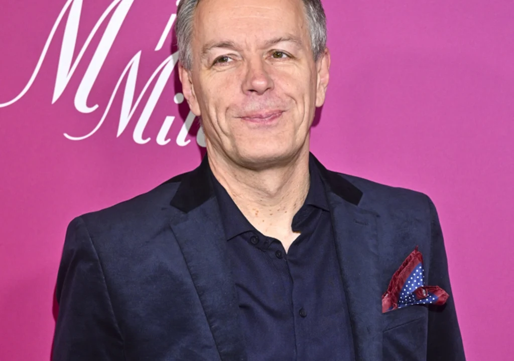 Mężczyzna w średnim wieku ubrany w granatową marynarkę oraz koszulę, z elegancko złożoną poszetką w kieszonce, stoi na tle różowej ścianki z białym fragmentem napisu.