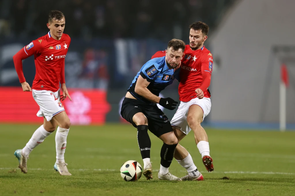 Trzech piłkarzy rywalizuje o piłkę podczas meczu na boisku, dwóch w czerwonych koszulkach próbuje odebrać piłkę zawodnikowi w ciemnoniebieskiej koszulce.