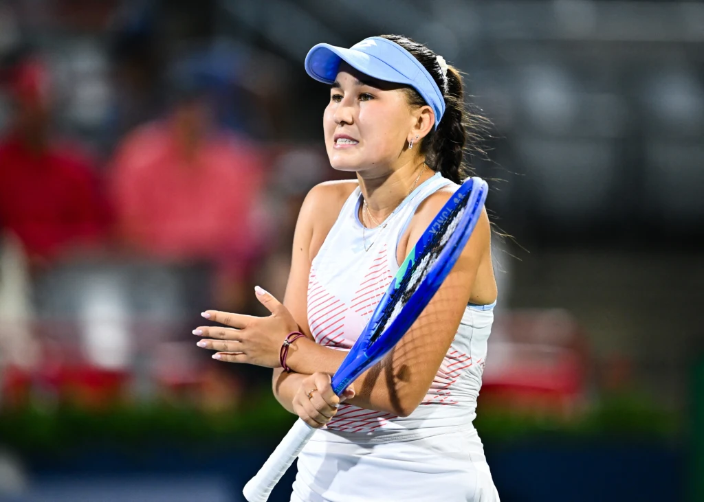 Tenisistka w białym stroju sportowym i niebieskiej czapce z daszkiem wyraża emocje podczas meczu, trzymając rakietę tenisową, w tle rozmazany obraz publiczności na trybunach.