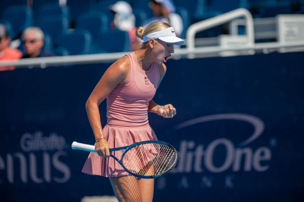 Tenisistka w sportowym stroju świętuje punkt na korcie podczas meczu tenisowego, w tle widoczne rozmyte sylwetki kibiców.