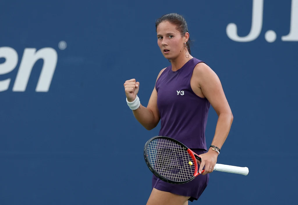 Tenisistka w fioletowym stroju sportowym na korcie, trzymająca rakietę w jednej ręce i zaciśniętą pięścią w geście triumfu, w tle niebieska ściana z fragmentem logo turnieju.