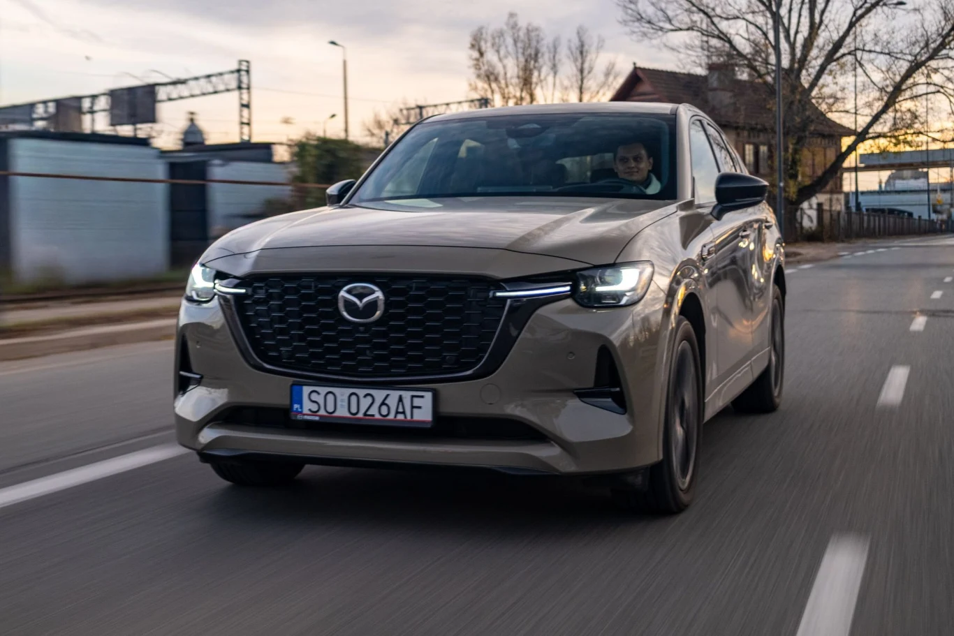 Jasnoszara Mazda SUV poruszająca się miejską ulicą o zmierzchu, wyraźnie widoczna przednia część auta z charakterystycznym grillem i reflektorami, na tablicy rejestracyjnej numer SO 026AF, w tle drzewa oraz zabudowania miejskie.