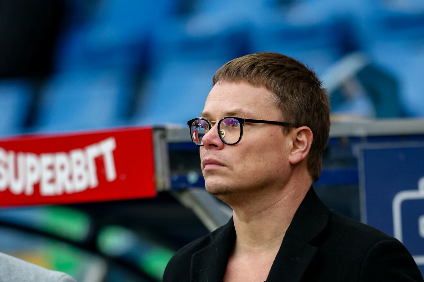 Jarosław Królewski Mężczyzna w okularach i czarnej marynarce patrzy przed siebie podczas wydarzenia sportowego, w tle rozmazane niebieskie trybuny stadionu i fragment czerwonego baneru reklamowego.