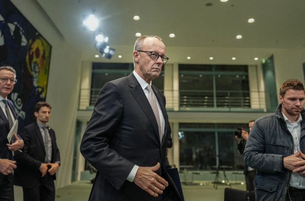 Kanclerz Niemiec Friedrich Merz ostrzega buntowników Mężczyzna w garniturze idący przez jasne, nowoczesne wnętrze budynku, wokół niego znajdują się inni ludzie w garniturach, a w tle widoczne są duże okna i reflektory.