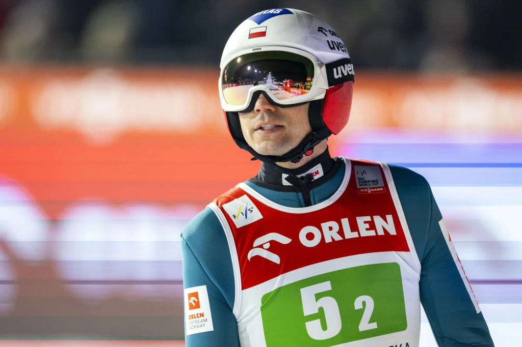 Kamil Stoch Skoczek narciarski w kombinezonie sportowym z numerem startowym 52 oraz kasku z flagą Polski, stojący na tle rozmytego stadionu podczas zawodów narciarskich. Na kamizelce widoczne logo sponsora Orlen.