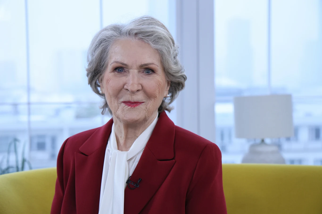 Odnaleziono nagrania Ireny Santor sprzed 50 lat Starsza kobieta z siwymi włosami w eleganckim bordowym żakiecie i białej apaszce siedzi na żółtej sofie w jasnym, nowoczesnym wnętrzu z dużymi oknami i lampą w tle. Uśmiecha się do aparatu.