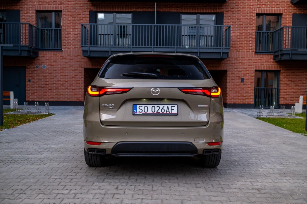Tylny widok beżowego samochodu Mazda CX-60 Hybrid zaparkowanego na brukowanym podjeździe, w tle nowoczesny budynek z czerwonej cegły z balkonami.