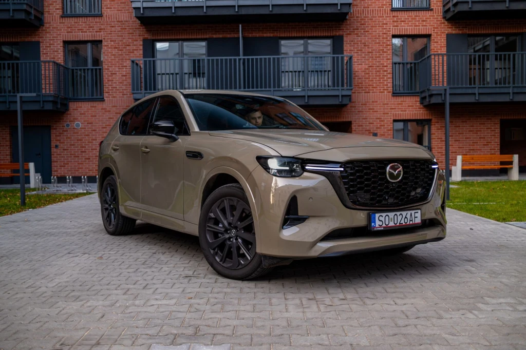 Beżowy samochód marki Mazda stoi zaparkowany na brukowanym podjeździe, w tle widać nowoczesny budynek z balkonami oraz zielony trawnik.