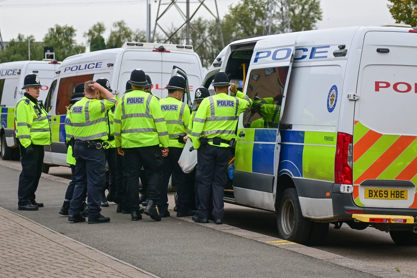 Grupa policjantów w jasnych kamizelkach odblaskowych stoi obok radiowozów policyjnych zaparkowanych przy ulicy. Funkcjonariusze najwyraźniej omawiają działania lub przygotowują się do akcji.