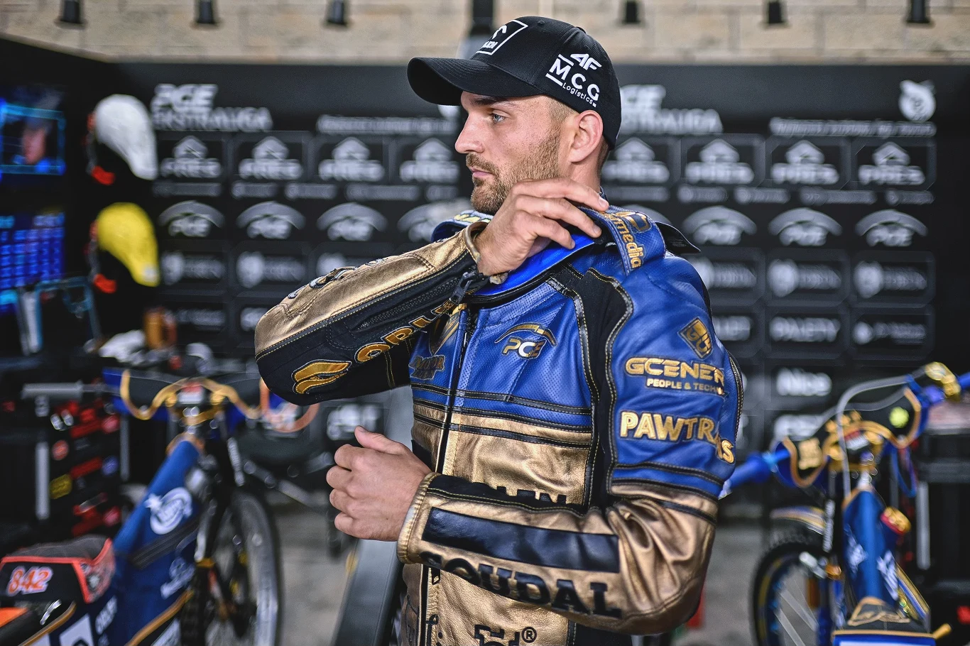 Żużlowiec w niebiesko-złoto-brązowym kombinezonie sportowym przygotowujący się do wyścigu na tle boksów z motocyklami oraz tablicami zespołu.