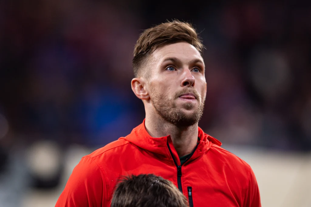 Mężczyzna w czerwonej bluzie sportowej stoi na tle rozmytego stadionu, patrząc lekko w górę, ze spokojnym wyrazem twarzy.