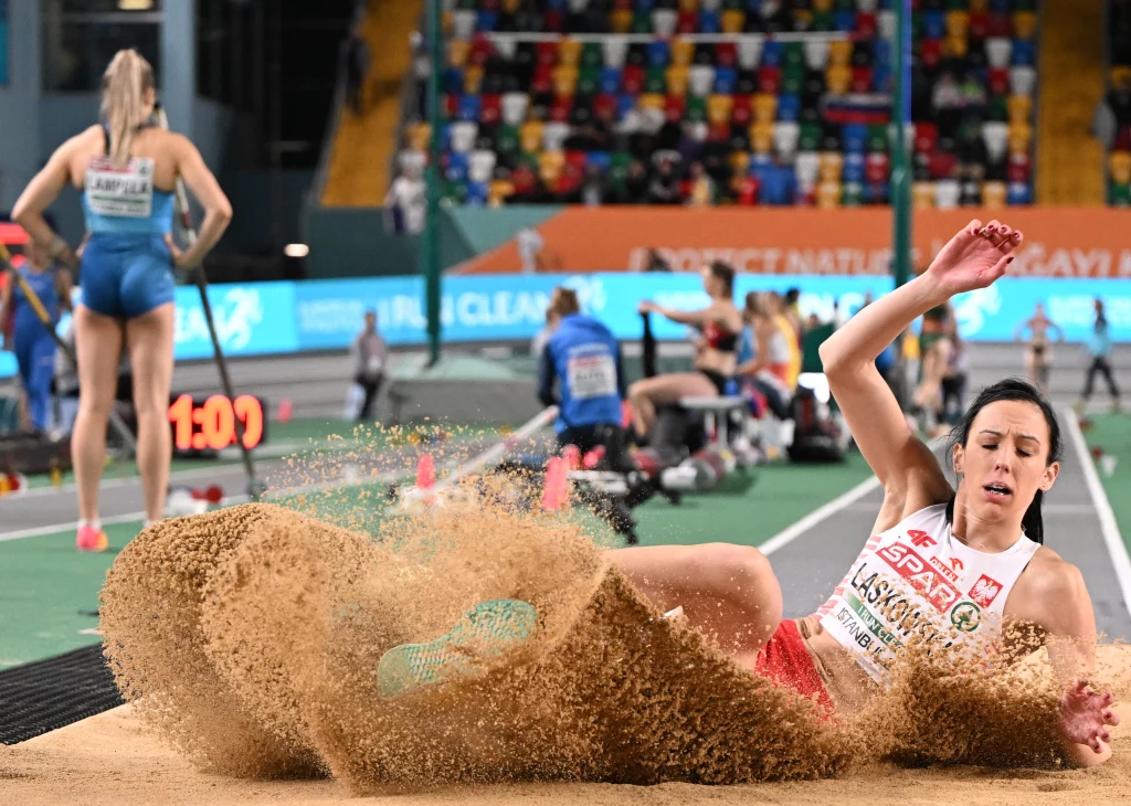 Zawodniczka wykonująca skok w dal ląduje w piaskownicy podczas zawodów lekkoatletycznych, w tle znajdują się inni sportowcy, trenerzy i kolorowe trybuny.