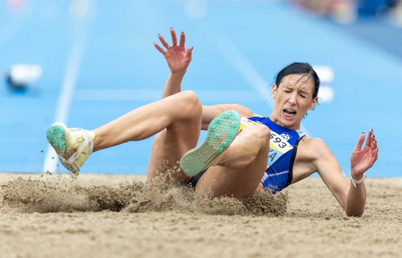 Zawodniczka lekkoatletyczna w trakcie lądowania w piaskownicy po skoku w dal, sportowy strój, napięcie mięśni i wyraz trudności na twarzy, niebieska bieżnia w tle.