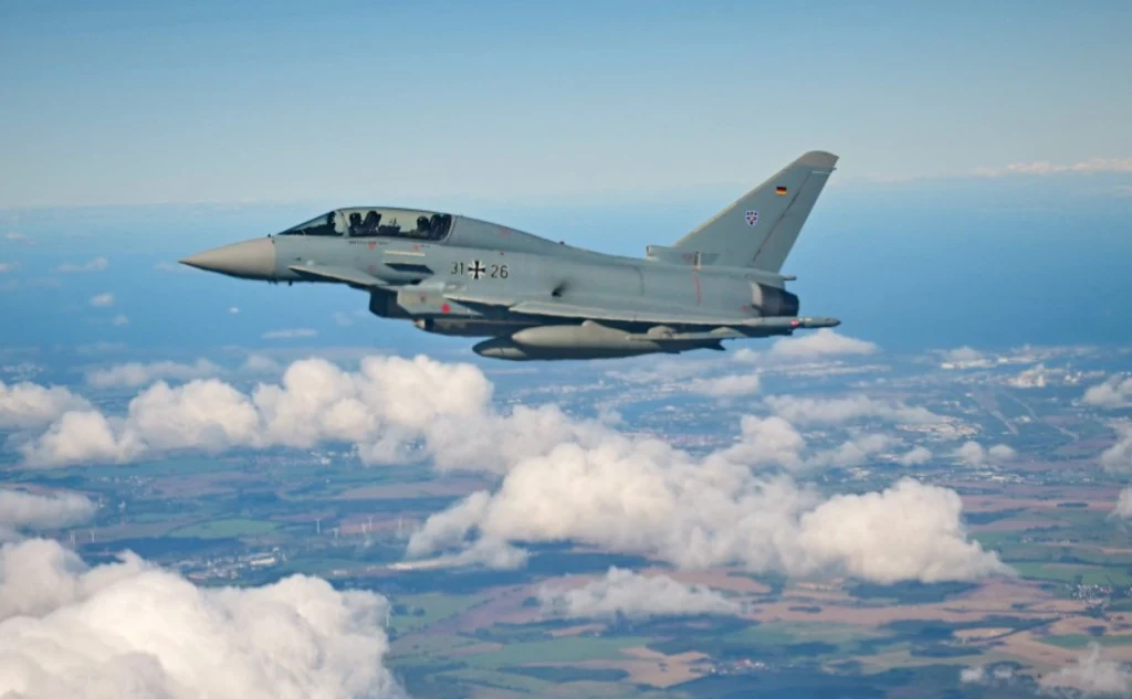 Myśliwiec wojskowy Eurofighter Typhoon z niemieckimi oznaczeniami przelatujący nad krajobrazem z rozproszonymi chmurami, w tle widać pola uprawne i fragmenty lasów.