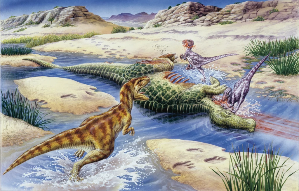 Nanotyrany przy padłym dinozaurze. Ten drapieżnik był trzy razy mniejszy niż t-rex Trzy drapieżne dinozaury rozszarpują ciało dużego gada leżącego w płytkim strumieniu, krew z ran miesza się z wodą, krajobraz uzupełniają skały i roślinność terenów pustynnych.