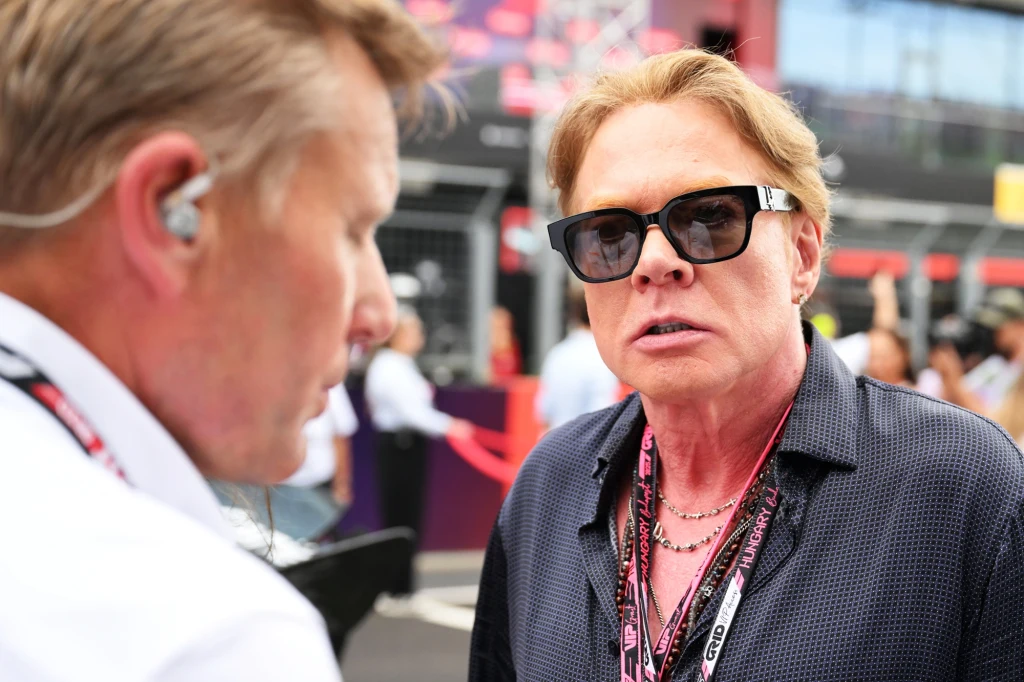 Dwóch mężczyzn rozmawia ze sobą na tle wydarzenia sportowego, jeden z nich nosi ciemne okulary, ma blond włosy i kilka naszyjników, drugi używa słuchawki na uchu.