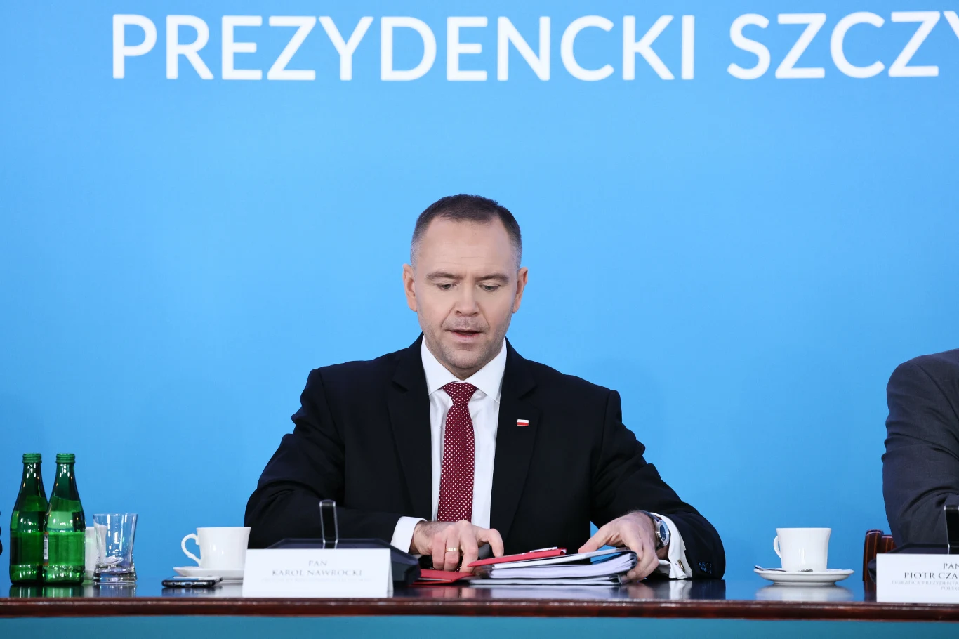 Karol Nawrocki zdecydował ws. Funduszu Medycznego Mężczyzna w garniturze siedzi przy stole na tle niebieskiej planszy z napisem, przed nim stos dokumentów, filiżanki, butelki wody i mikrofon.