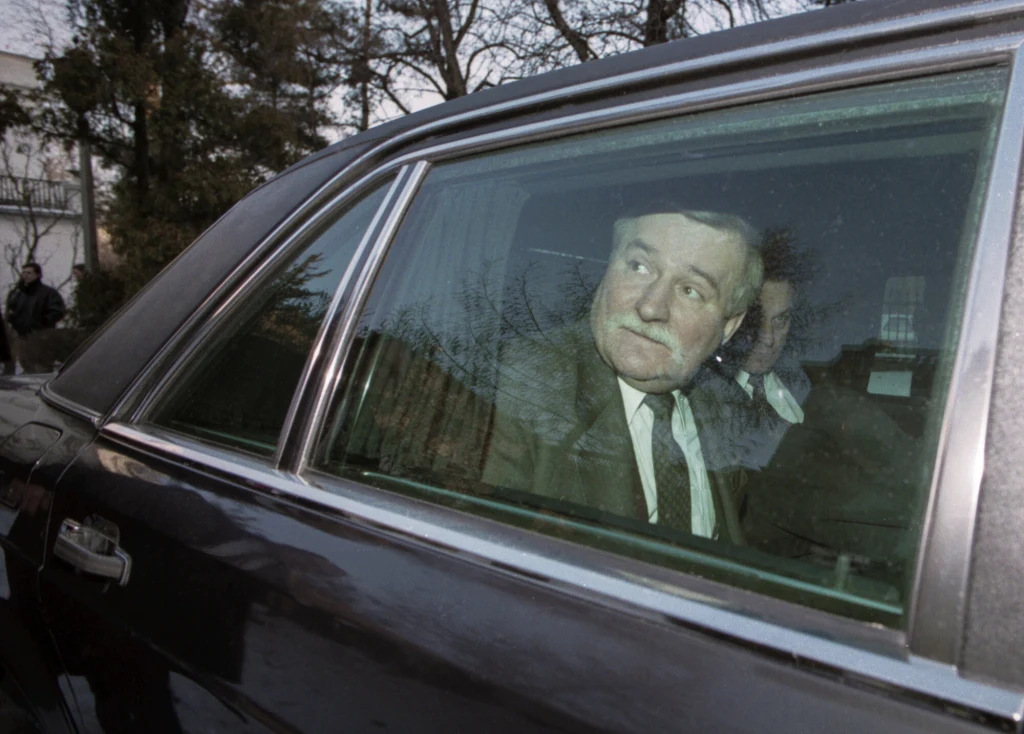 Były prezydent RP Lech Wałęsa Mężczyzna w marynarce i krawacie siedzi na tylnym siedzeniu samochodu, patrzy przez szybę na zewnątrz. W tle widoczne są drzewa i fragmenty budynków.
