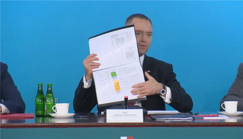 Mężczyzna w garniturze prezentuje otwarty segregator lub dokument na tle niebieskiej ściany. Przed nim na stole stoją dwie zielone butelki, filiżanka z kawą oraz tabliczka z imieniem i nazwiskiem. Po obu stronach widoczne są fragmenty rąk innych osób s...