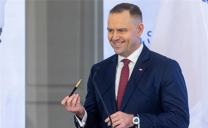 Mężczyzna w garniturze z czerwonym krawatem stoi przy mikrofonie, trzyma długopis w prawej ręce i uśmiecha się. W tle widoczna biała flaga i jasna ściana.