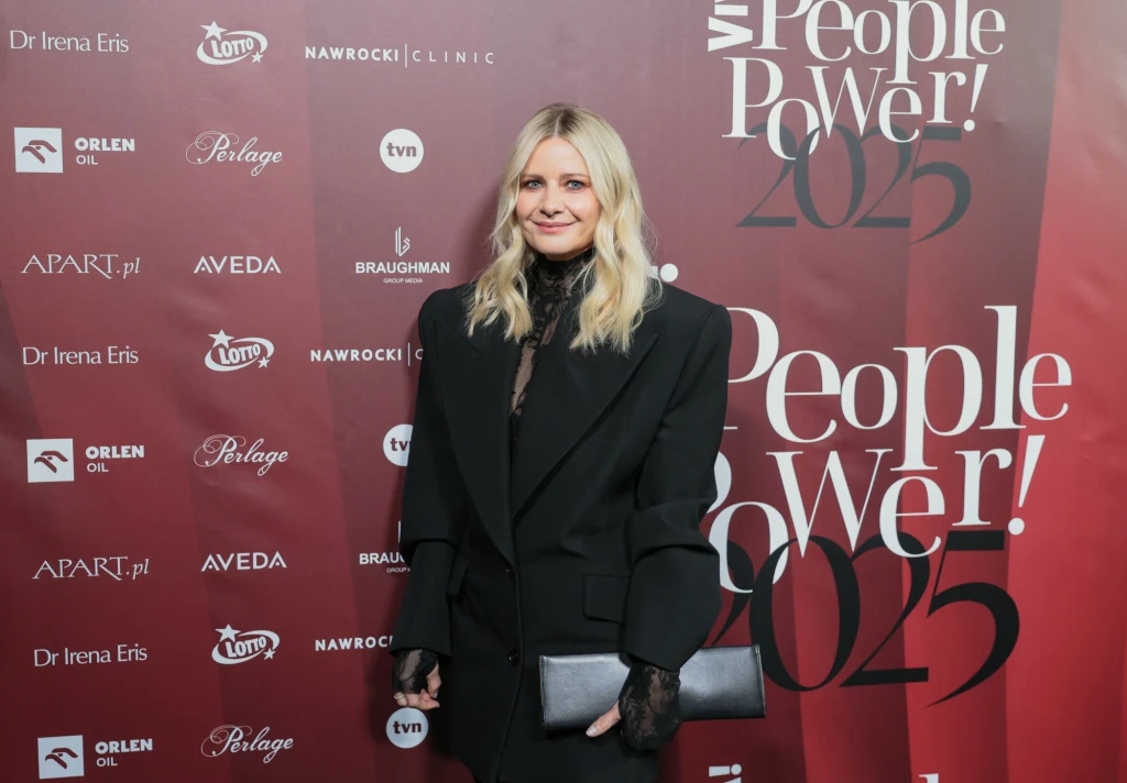 Kobieta w eleganckim czarnym garniturze stoi z torebką w ręku na tle ścianki z logotypami sponsorów oraz napisem People Power 2025.