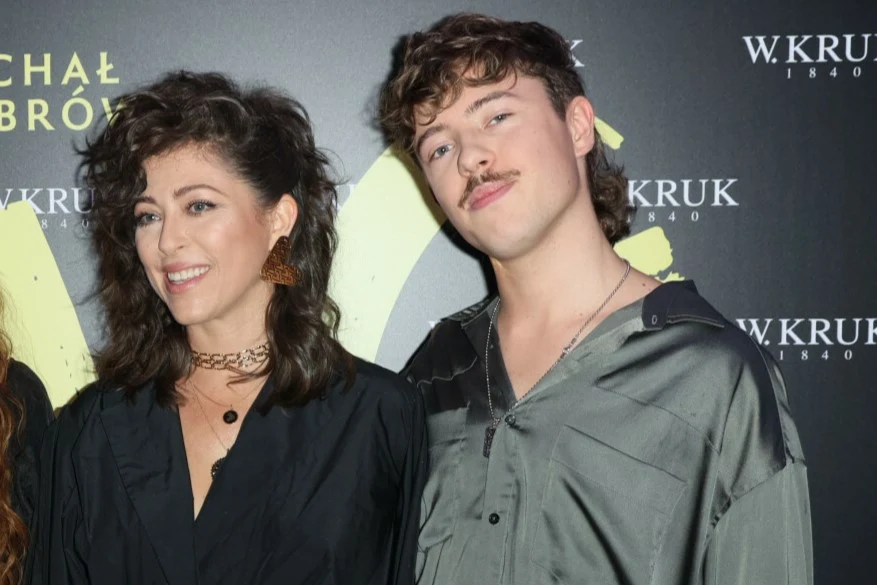 Dwie uśmiechnięte osoby pozujące razem podczas eventu, jedna z nich ma na sobie czarną koszulę i duże kolczyki, druga jasnoszarą koszulę z rozpiętym kołnierzykiem, w tle widoczne logo marki biżuteryjnej.