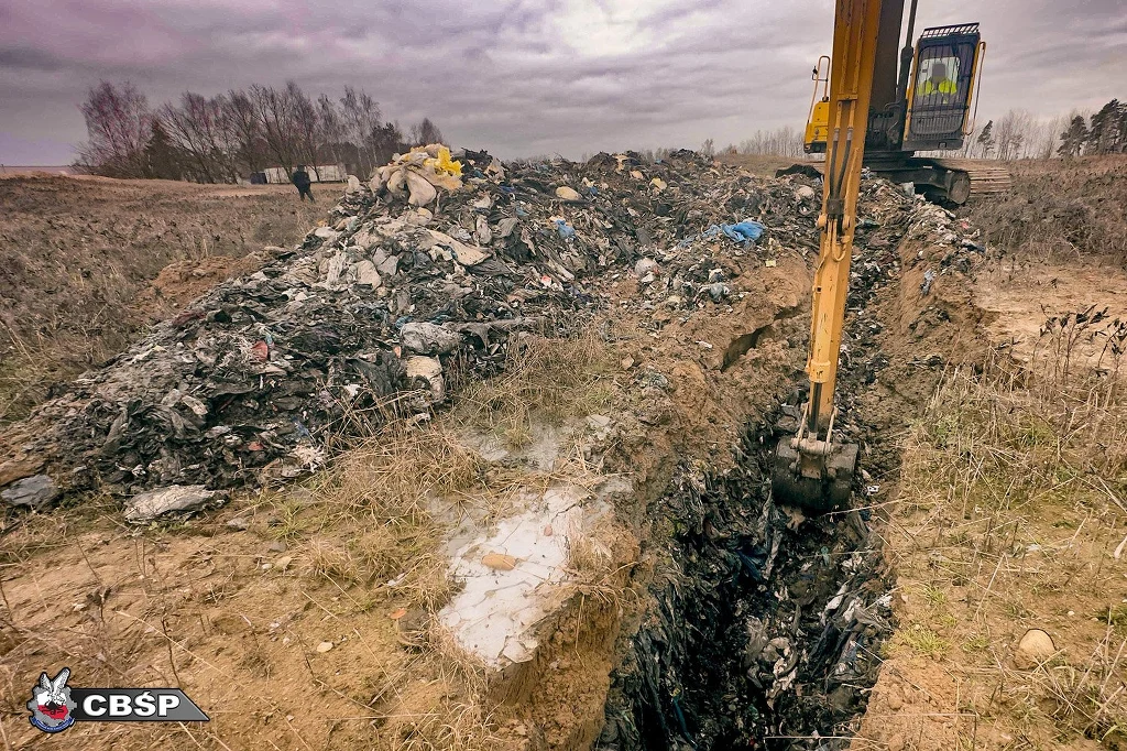 Koparka zakopuje dużą ilość odpadów na otwartym terenie, wokół znajdują się stosy śmieci i rozległy krajobraz z drzewami i chmurami na niebie.