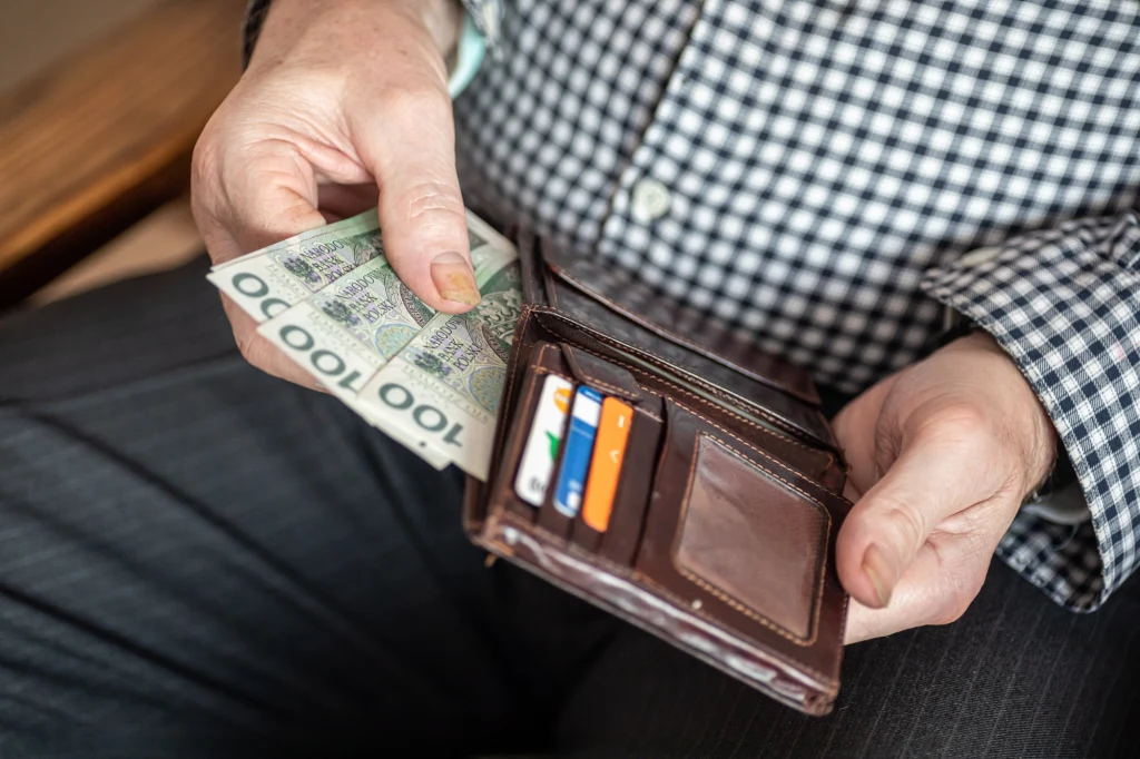 Ta grupa emerytów otrzyma wyższe świadczenia. Przeliczenie nastąpi automatycznie Portfel trzymany w dłoniach mężczyzny w koszuli w kratkę z widocznymi banknotami 100 złotych oraz kartami płatniczymi.