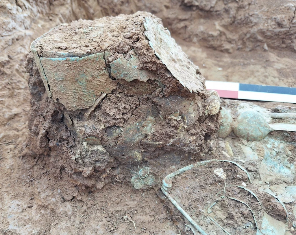 Dama z odwróconą koroną. Tajemniczy grób młodej arystokratki odkryty w Grecji Fragment wykopalisk archeologicznych ukazujący ziemię oraz odkryte metalowe i gliniane artefakty, częściowo zniszczone i pokryte ziemią, w towarzystwie narzędzia pomiarowego.