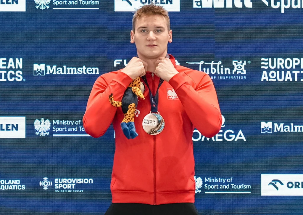 Kamil Sieradzki Sportowiec w czerwonej bluzie z medalem na szyi stoi przed ścianką sponsorską, trzymając maskotkę, tło zawiera loga partnerów i organizatorów wydarzenia.