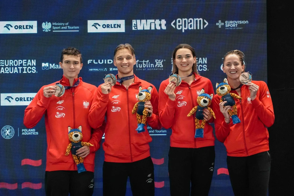 Aleksander Styś, Jan Kałusowski, Kornelia Fiedkiewicz, Katarzyna Wasick Czwórka młodych sportowców w czerwonych dresach narodowych trzyma medale oraz pluszowe maskotki, stojąc wspólnie na tle ścianki z logotypami partnerów i organizacji sportowych.