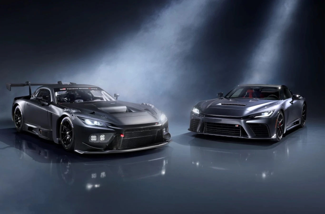 Światowa premiera Toyoty GR GT i GR GT3. Dwa czarne sportowe samochody stojące obok siebie pod padającym z góry światłem, jeden z wyraźnym pakietem aerodynamicznym i dużym spoilerem, drugi o bardziej cywilnym, eleganckim wyglądzie.