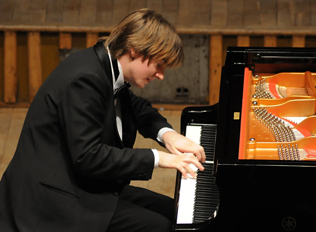 Młody mężczyzna w eleganckim garniturze gra na fortepianie koncertowym, skupiony na klawiaturze, otoczony wnętrzem sali koncertowej z drewnianą podłogą.