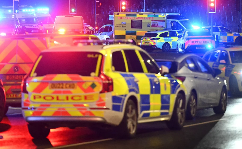 Kilka radiowozów policyjnych i karetki pogotowia z włączonymi światłami ostrzegawczymi na ruchliwej ulicy nocą, otoczonych przez inne pojazdy, w tle sygnalizacja świetlna.