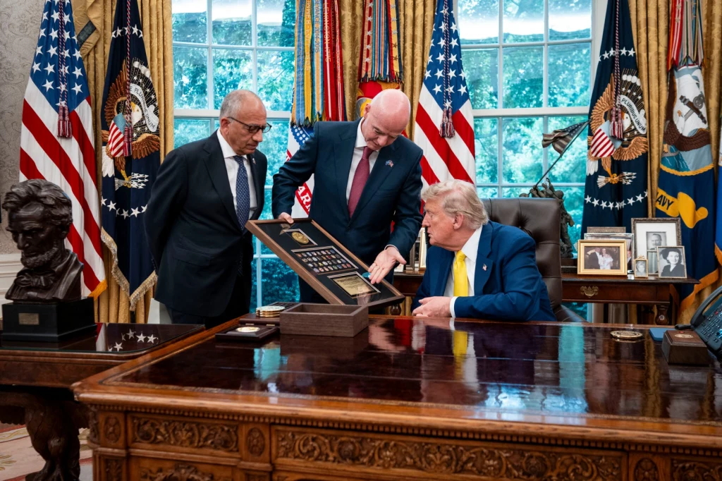 Donald Trump lubi otrzymywać prezenty Trzech mężczyzn ujętych w gabinecie o wystroju oficjalnym, jeden z nich siedzi za dużym biurkiem i jest ubrany w niebieski garnitur z żółtym krawatem, dwaj stoją obok i prezentują dużą tablicę z medalami lub odznaczeniami. Za nimi widać amerykańskie fl...