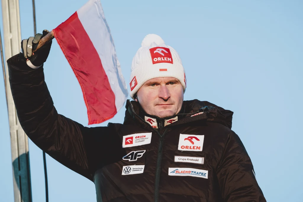 Maciej Maciusiak Mężczyzna w czapce i zimowej kurtce z licznymi logotypami sponsorów, trzymający w górze polską flagę na tle jasnego, niebieskiego nieba